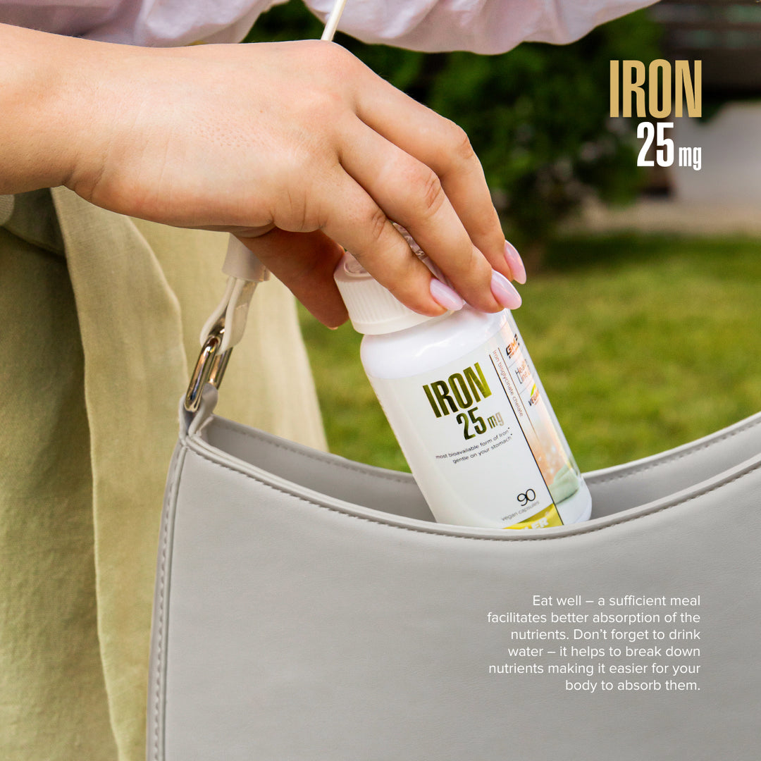 Person placing a supplement bottle labeled 'IRON 25 mg' into a handbag with a green outdoor background.